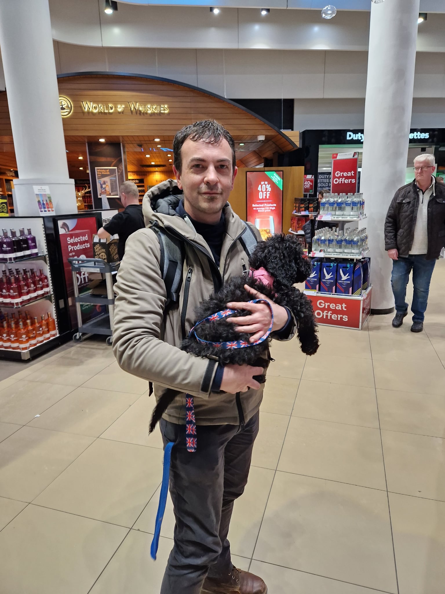 Andreas und Levi: Mann mit Rucksack hält einen schwarzen Welpen mit lockigem Fell im Arm, im Hintergrund ein Flughafen-Shop